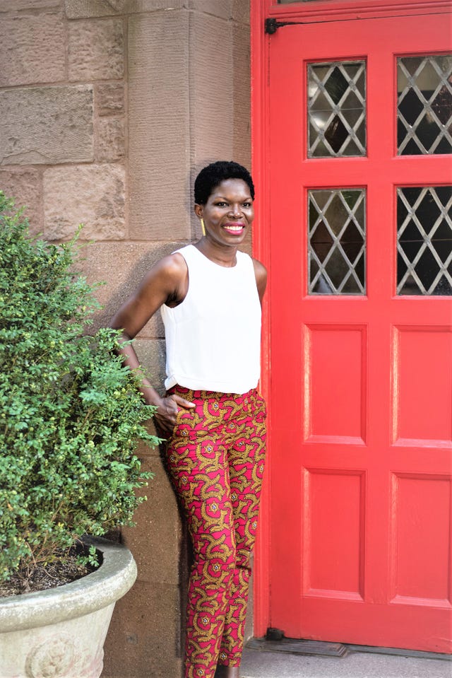 54kibo, woman with a white top and patterned pants next to a red door outdoors