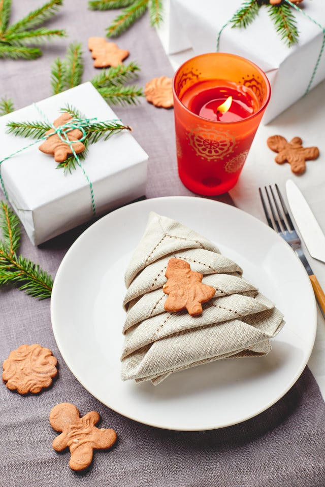 napkin in the form of a christmas tree on a plate on white tablecloth with gifts and decorations with fir sprigs and gingerbread cookies