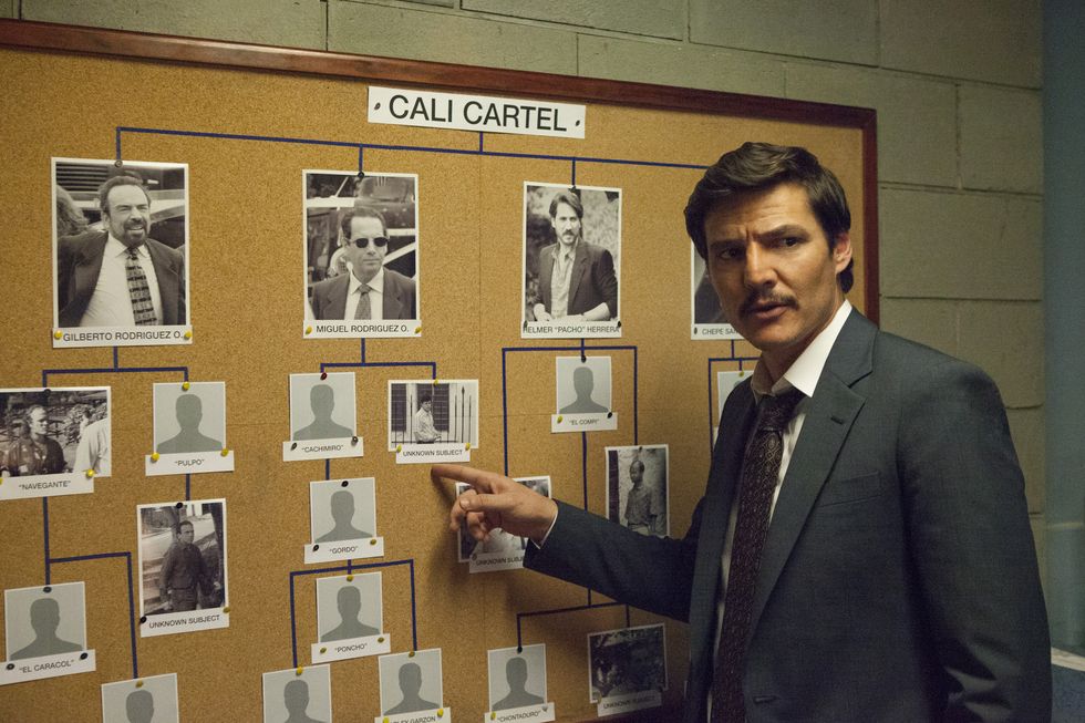 pedro pascal in character for narcos, he stands in front of and points at a cork board with a web of mugshots, he wears a gray suit