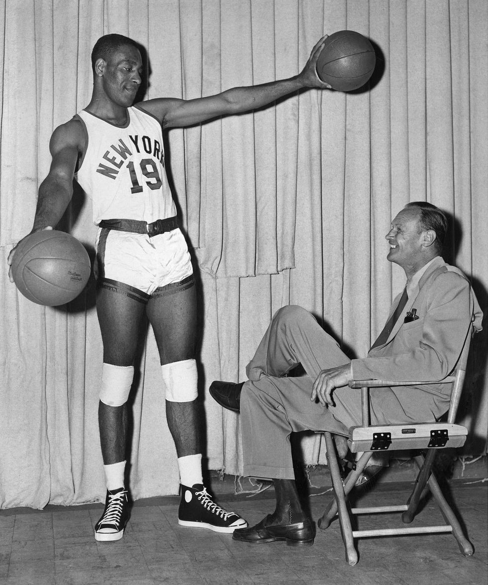 nat clifton holding two basketballs while wearing a new york knicks jersey, standing next to a seated joe lapchick