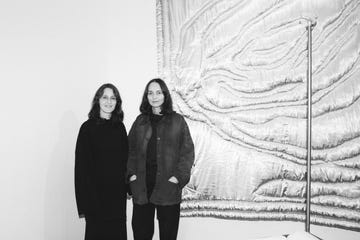 two individuals standing in front of a textured wall hanging