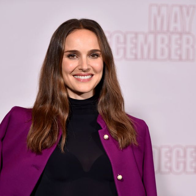natalie portman smiles at the camera, she wears a black turtleneck top and a violet jacket on her shoulders, she stands in front of a light background