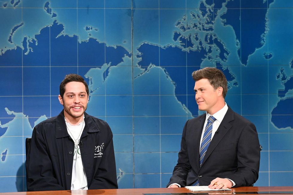 pete davidson, wearing a black jacket and white t shirt, sitting at a news desk next to colin jost in a black suit and blue tie, with a map of the world on the wall behind them
