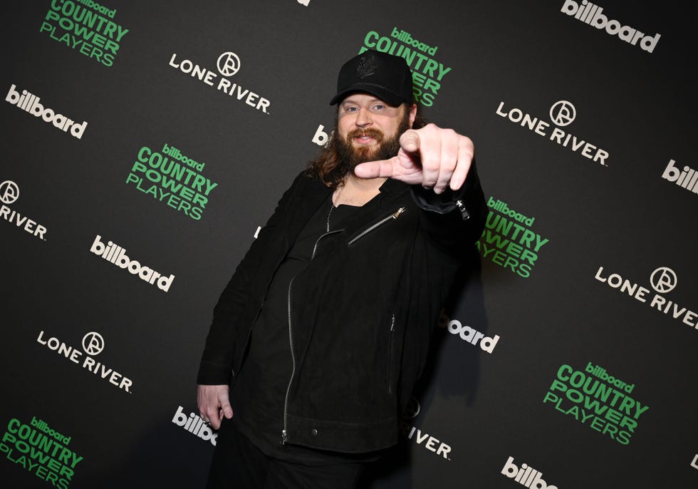 nate smith smiles and points at the camera, he stands in front of a black photo backdrop with logos on it and wears a black cap, jacket and pants