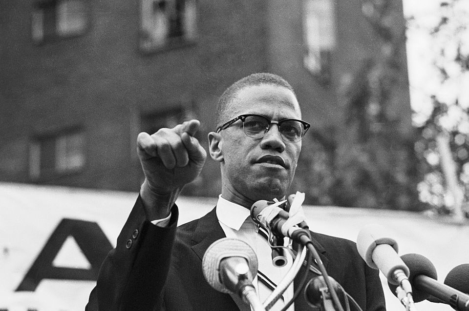 malcolm x speaking into several microphones and points with one finger as he stands outside, he wears glasses, a suit jacket and collared shirt