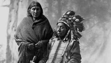 a woman stands next to red cloud who sits while wearing a feather headdress