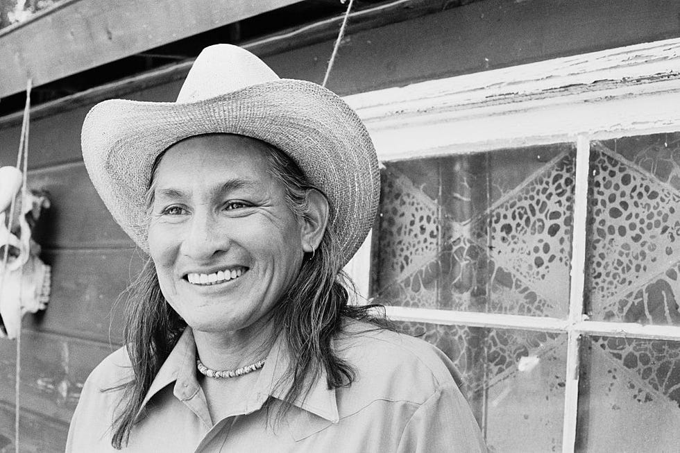 will sampson smiles and looks to the left, he wears a straw cowboy hat, collared shirt, and beaded chocker necklace
