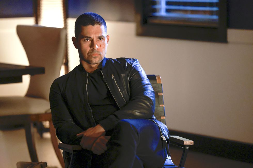 wilmer valderrama as nick torres in ncis season 20, a man sits in a chair looking stern, he wears a black tshirt, jeans and leather jacket