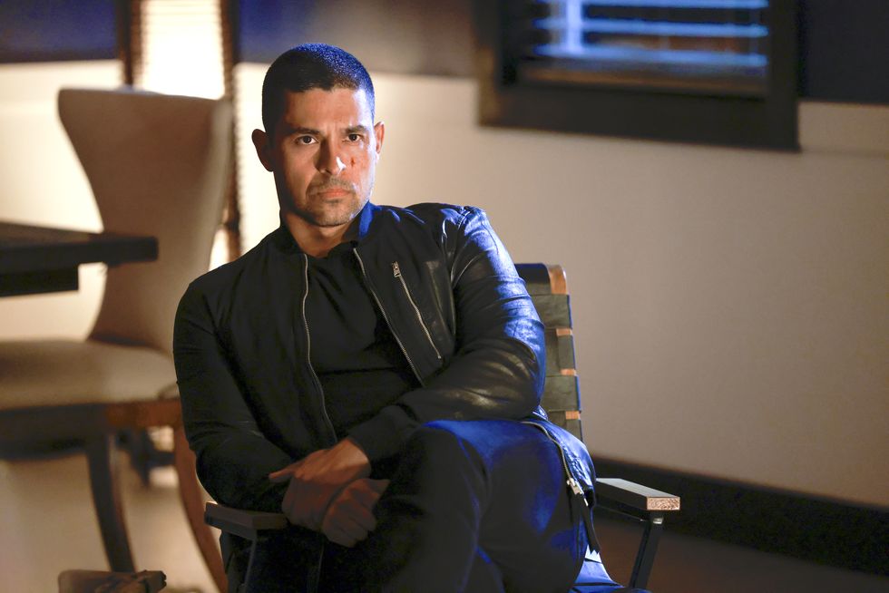 Wilmer Valderrama como Nick Torres en la temporada 20 de NCIS, un hombre sentado en una silla con aspecto severo, viste una camiseta negra, jeans y una chaqueta de cuero.