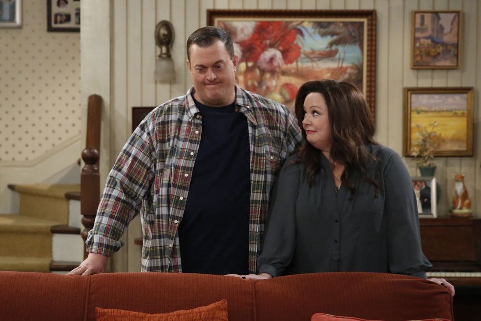 billy gardell and melissa mccarthy stand behind a couch and rest their hands against the back, gardell looks down and mccarthy looks at him