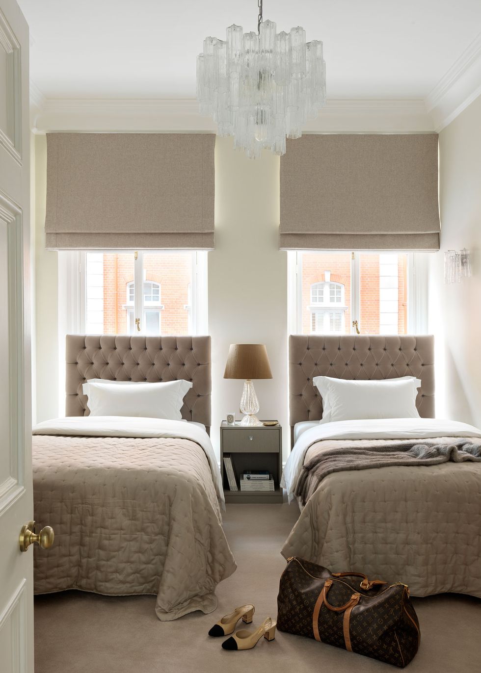 twin beds in a small bedroom with tufted headboards and tall windows behind them and in the foreground on a taupe rug there are a pair of sling backs and a lv travel bag
