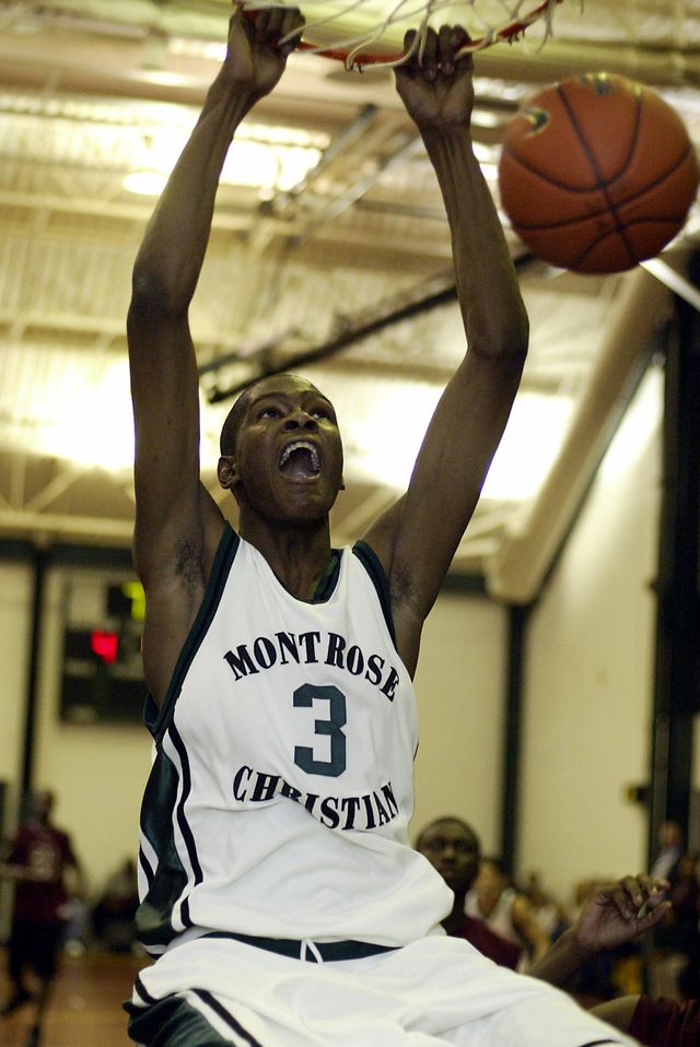 a teenage boy hangs from a basketball hoop after dunking the ball through, he has a wide open smile on his face and wears a mostly white basketball uniform