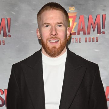 Neil Jones neil jones, a man stands with his hands behind his back looking at the camera with a slight smile, he has short ginger hair and beard, he wears a white jumper with black trousers and coat