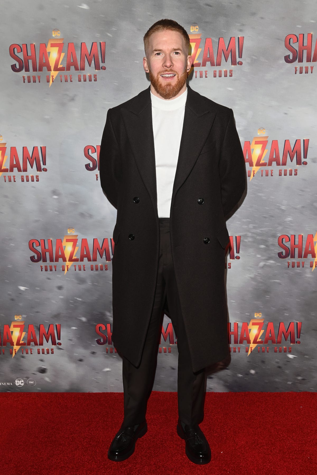 neil jones, a man stands with his hands behind his back looking at the camera with a slight smile, he has short ginger hair and beard, he wears a white jumper with black trousers and coat