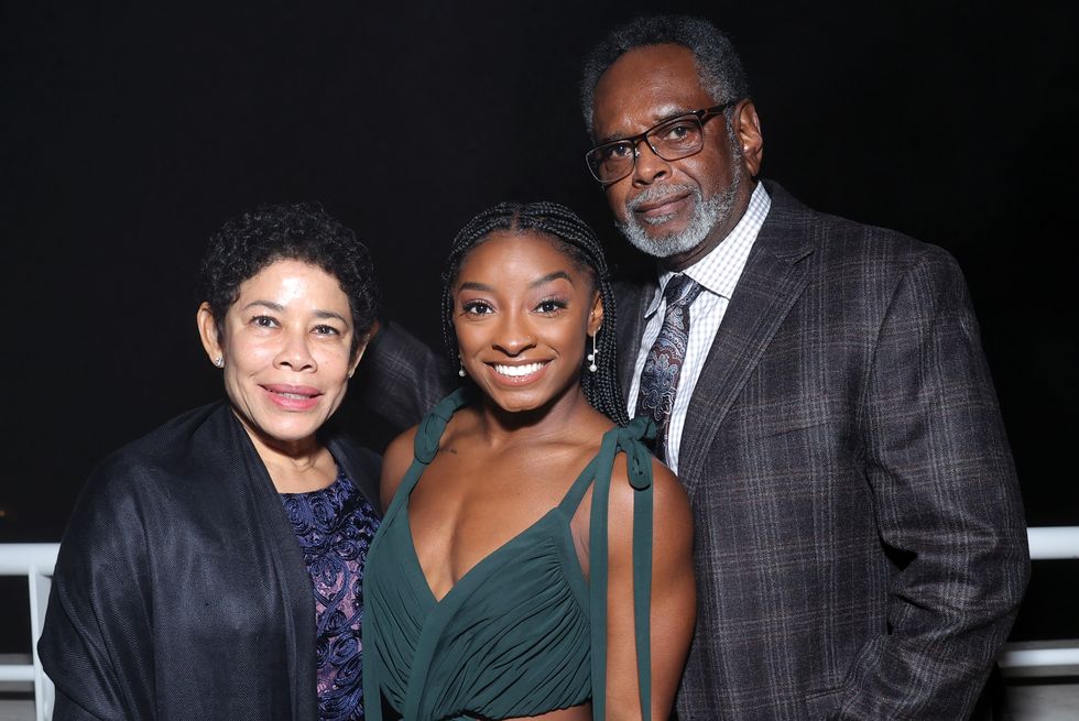 nellie biles, simone biles, and ron biles stand next to each other and embrace for a photo, all three smile for the camera and wear formal clothing