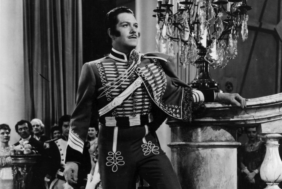 Nelson Eddy in “Phantom Of The Opera” a black and white photo of nelson eddy on a movie set, wearing an aristocratic period costume, looking up from the bottom of a staircase, with several extras standing behind him