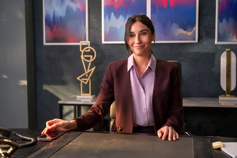 Lilah Richcreek Estrada sits at a desk as Julie and smiles in a scene from the Netflix sitcom A Man on the Inside