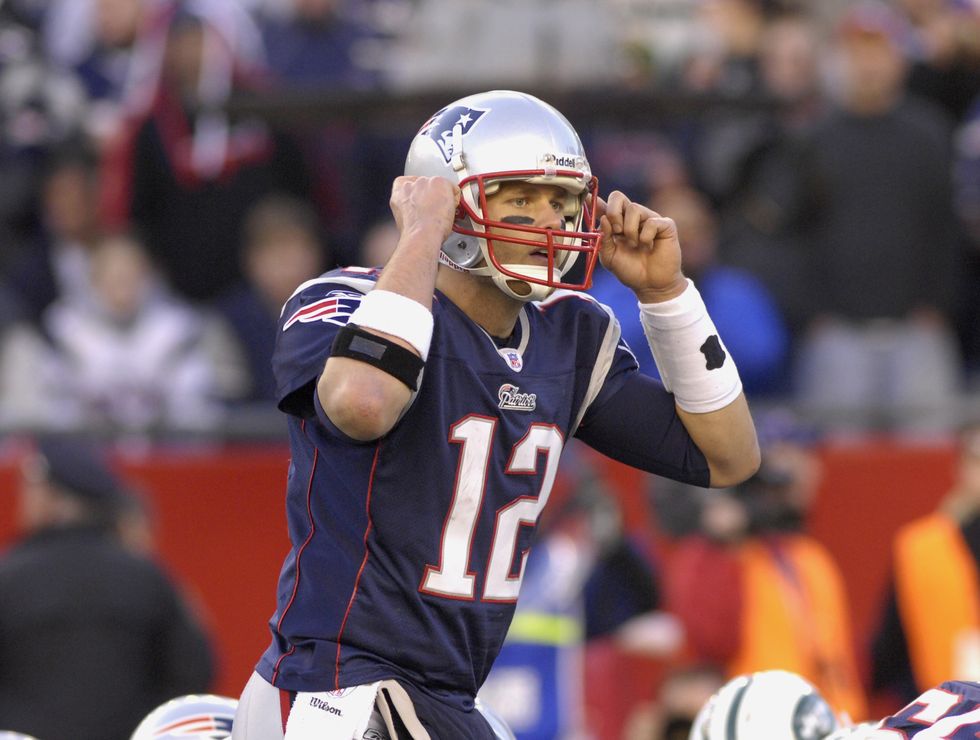 tom brady wearing a full football uniform and raising his hands toward both sides of his helmet on the field