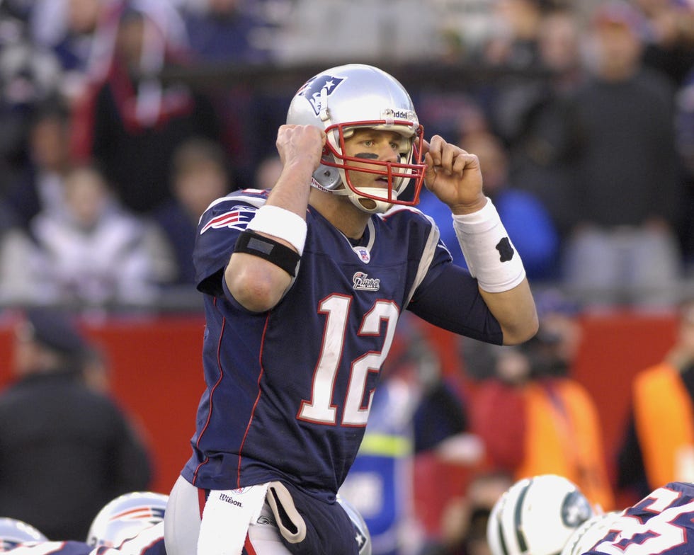 tom brady wearing a full football uniform and raising his hands toward both sides of his helmet on the field