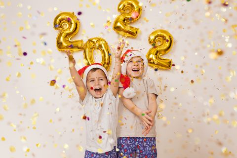 happy smiling kids in red santa hat are enjoying confetti near white wall with gold balloons celebrating new year 2022 at home christmas concept