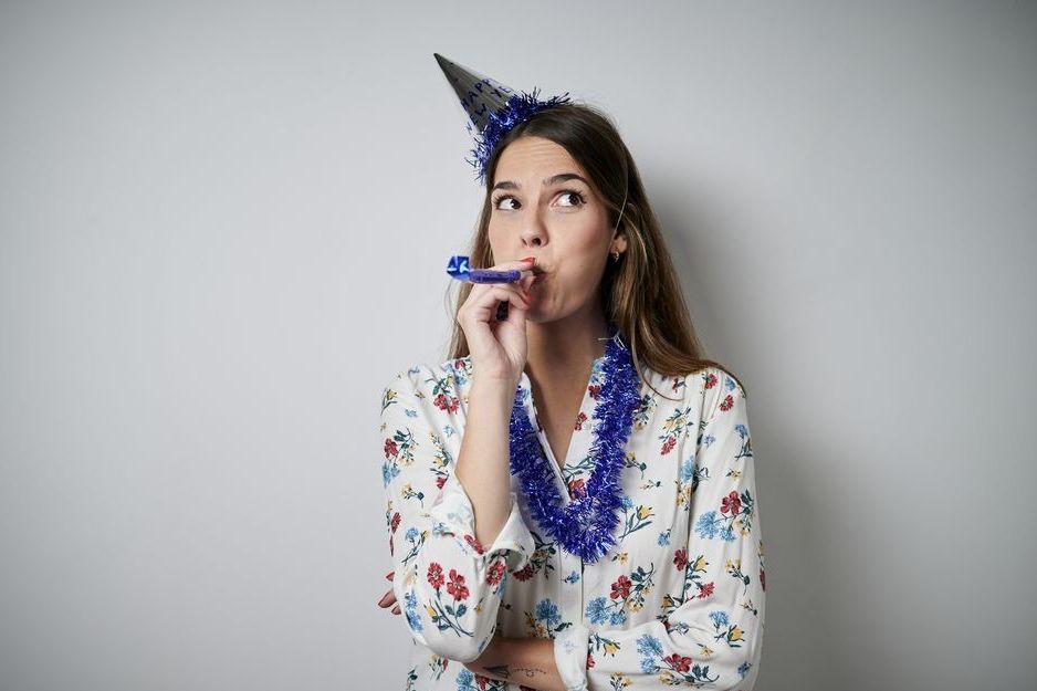 woman in party hat ironically blowing a party horn with a bored look on her face, possibly on new year's