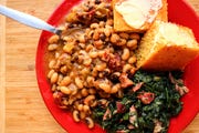 stewed black eyed peas on a red plate, alongside greens and buttered cornbread, all on a wooden surface