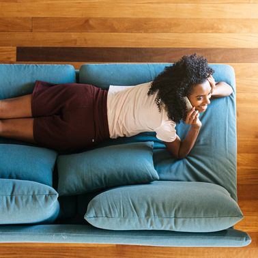 top view of african american lady in casual wear resting on blue sofa and smiling while surfing on mobile phone at home