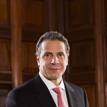 a man in a black suit jacket white collared shirt and red patterned tie smiles while standing in front of a wooden paneled wall