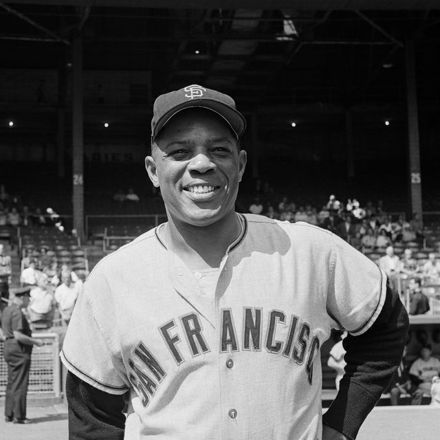 willie mays stands on a baseball diamond and smiles, he wears a baseball hat and uniform for the san francisco giants