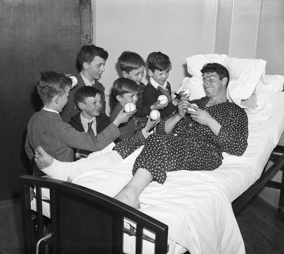 joe dimaggio lies in a hospital bed with a wrap around one leg and foot, he holds a baseball and marker as several boys stand to the left of the be smiling and waiting for autographs