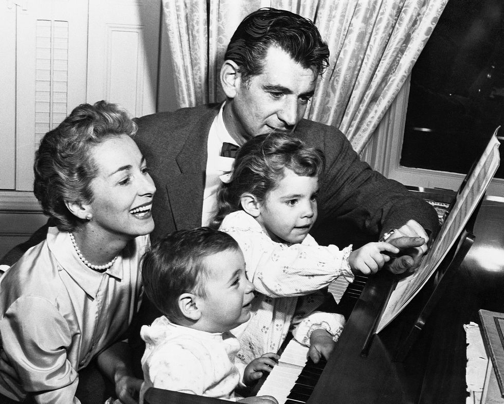 leonard bernstein sits at a piano with his two young children, felicia montealegre sits behind the kids and smiles, she wears a pearl necklace and satin collared shirt, bernstein has on a suit