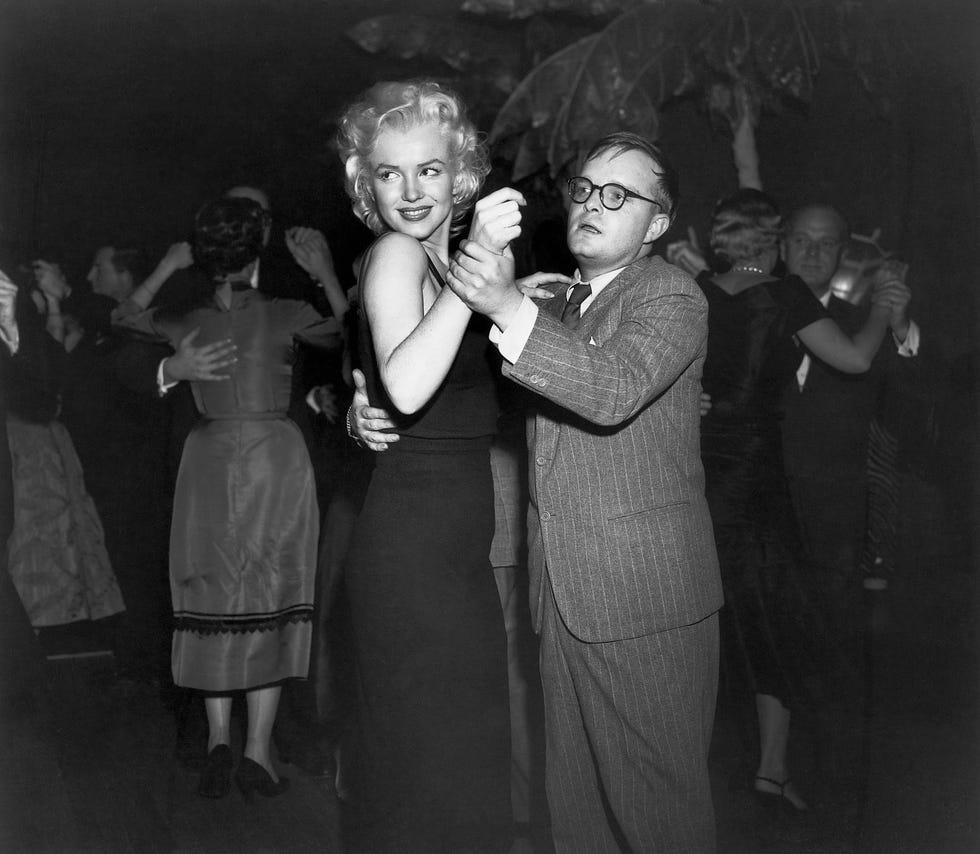 marilyn monroe and truman capote dance with other couples dancing in the background, she wears a sleeveless dress and smiles, he wears a suit with a tie and glasses