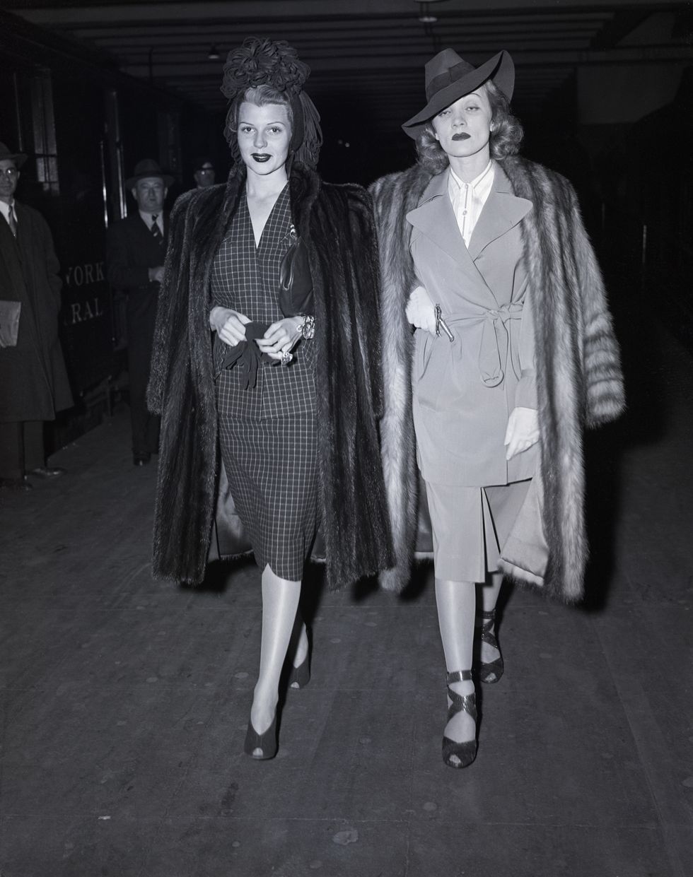 rita hayworth and marlene dietrich walk together, both wear fur jackets on their shoulders, hats, skirt suits and high heels