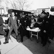 police roll a stretcher with malcolm x body on a sidewalk, many people are on the street as well as a police truck and cars