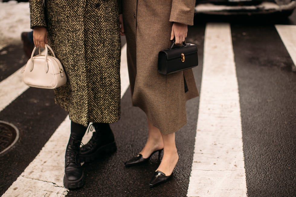 two individuals standing on a crosswalk displaying their fashionable outfits and handbags