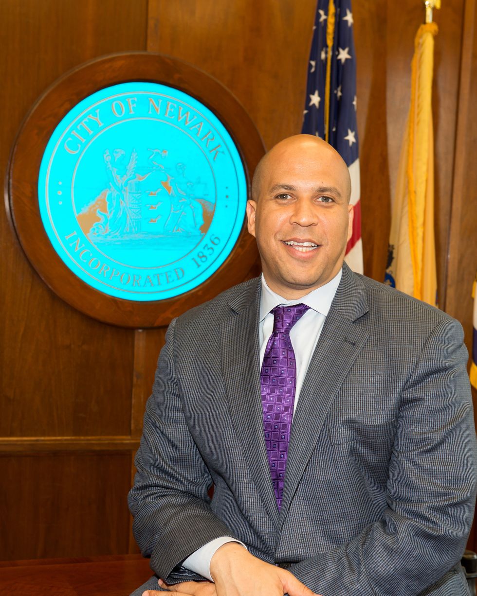 a bald man smiles at the camera inside a wood paneled room with a large blue emblem, he wears a gray checked suit jacket collared shirt and purple tie