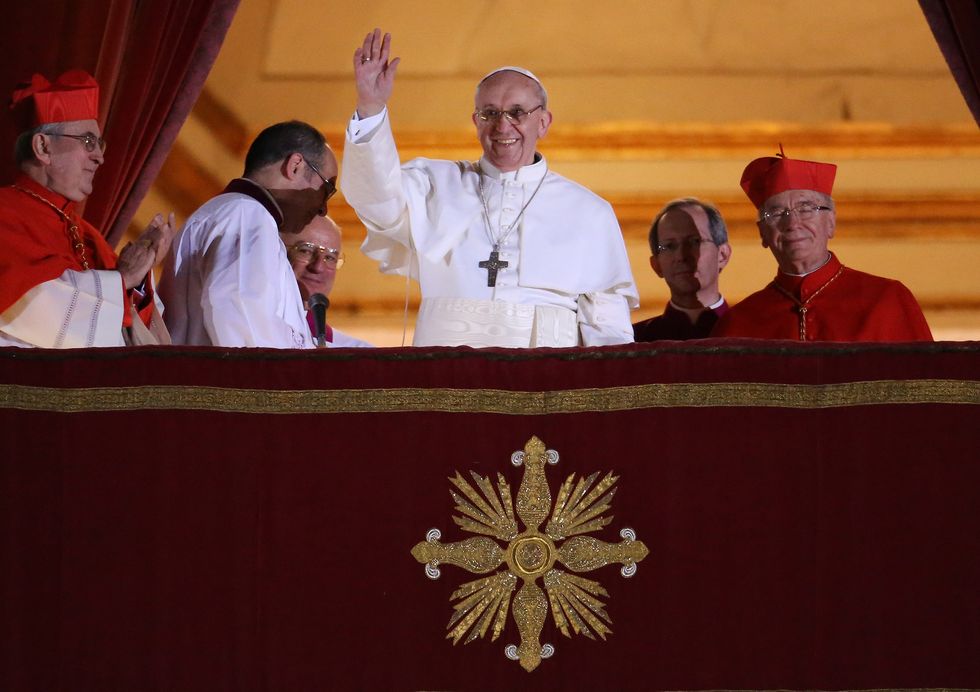 pope francis stands on a balcony and waves as several men stand on either side of him, all wear priest robes