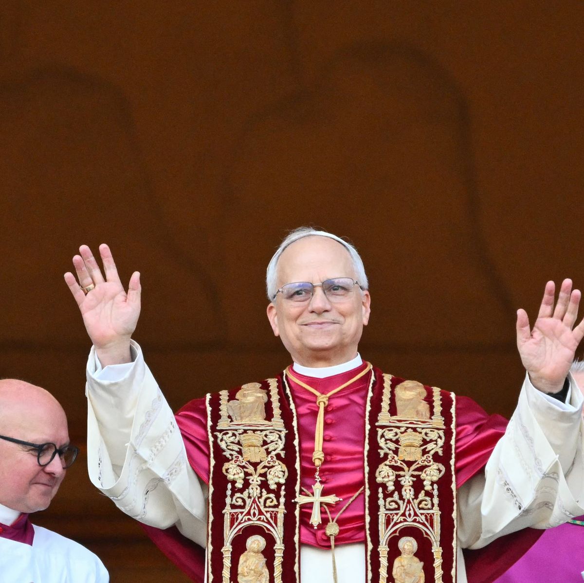a man in white and red robes smiles and waves from a balcony, two men stand behind him on either side