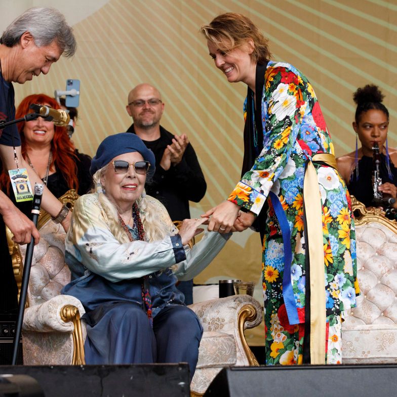joni mitchell smiles while sitting in an armchair, she holds brandi carliles hands who stands to the right, mitchell wears a blue beret, large sunglasses, and a blue and white outfit, other people are also on the stage in the background