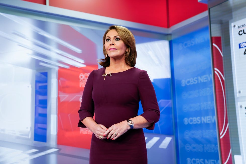 maria elena salinas stands on a cbs news television set with red, white, and blue backgrounds, she wears a purple long sleeve dress with a watch and hoop earrings