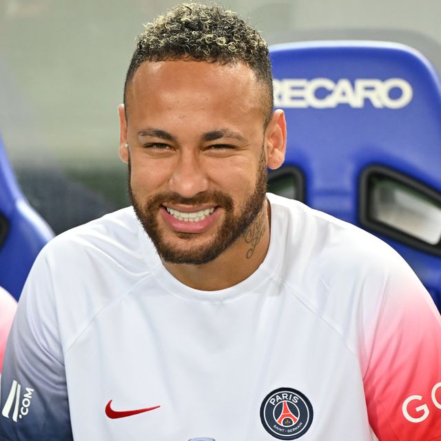 neymar smiles at the camera while sitting in a blue chair and wearing a white paris saint germain jersey with one blue sleeve and one red sleeve