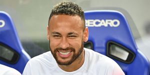 neymar smiles at the camera while sitting in a blue chair and wearing a white paris saint germain jersey with one blue sleeve and one red sleeve