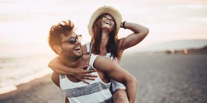 man in striped tank and sunglasses giving woman in straw hat a piggyback ride on a beach they are laughing
