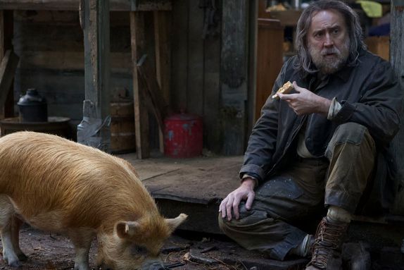 nicolas cage in pig trailer sitting down outside a cabin next to a pig