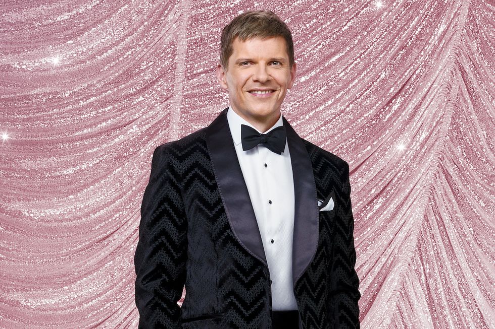 Nigel Harman w Strictly Come Dancing 2023, mężczyzna stojący z rękami w kieszeniach, patrzący w kamerę i uśmiechnięty, ubrany w czarny garnitur z krawatem i otoczony błyszczącymi kulkami