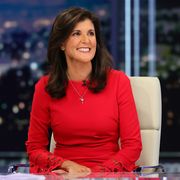 nikki haley sits in a white tall backed desk chair and smiles while looking to the right side of the frame, she is wearing a bright red long sleeve dress that has red lace detailing at the sleeve cuffs and a silver pendant necklace, her brown hair is styled down with loose curls