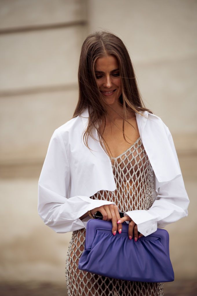 a woman wearing a net dress, cropped white shirt, and purple leather clutch on the street during copenhagen fashion week spring summer 2023 in a roundup of cute summer outfits 2023
