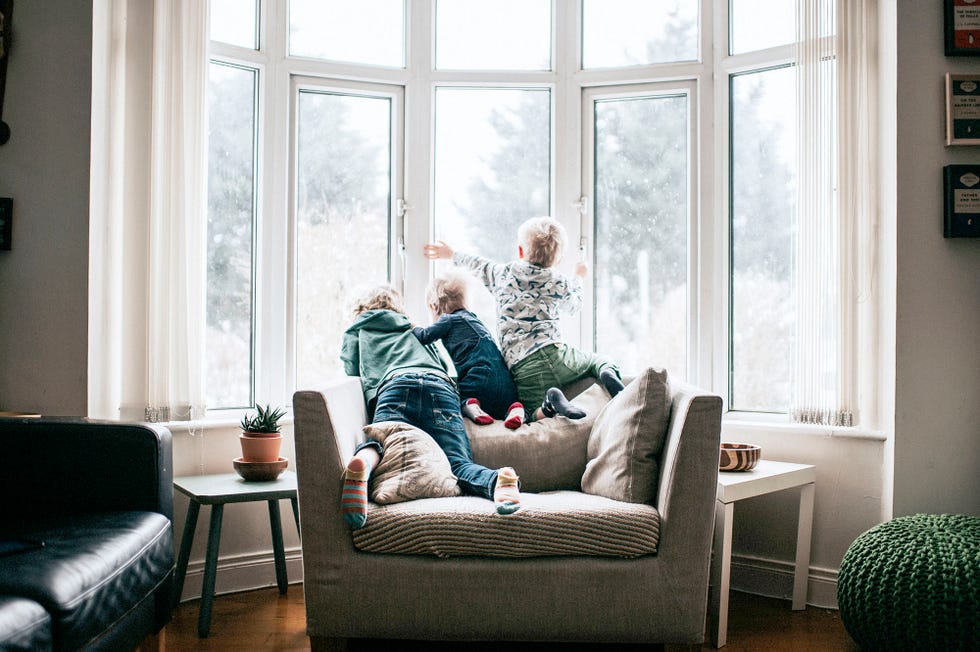 tres ninos subidos a un sillon miran por la ventana de su casa mientras fuera nieva