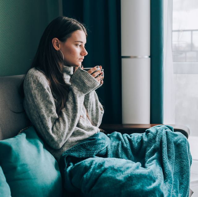 no central heating, cold apartment young brunette woman drink hot tea, look out the window, wrapped in a blanket in cold apartment