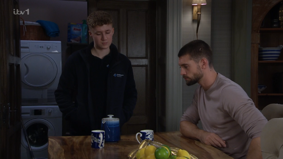 noah dingle stands by a table, joe is sat down with two cups of tea in front of him on a scene of emmerdale